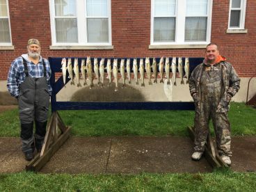 Two anglers proudly posing beside a catch board displaying a variety of Walleye after a successful fishing trip.