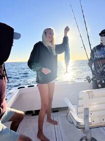 A young woman joyfully displays her catch, a fish, while fishing on a boat during a sunny day at sea.