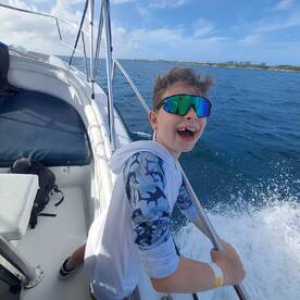A young boy enjoying the thrill of a fishing trip aboard a boat, smiling widely as he holds onto the railing with the ocean in the background.