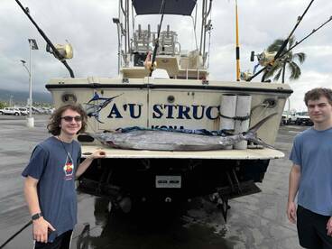 Two young anglers proudly displaying a large fish on the back of a boat named 'A'U Struck' at a marina, showcasing their successful fishing trip.