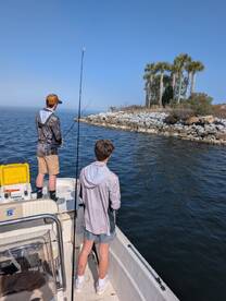 Two young anglers casting lines from a boat near a rocky shoreline, surrounded by palm trees on a clear day.