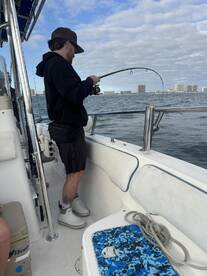 An angler is actively reeling in a fish while fishing from a boat, with a scenic backdrop of buildings along the waterfront.