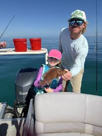 A young girl proudly displaying a colorful Grouper while fishing on a boat, accompanied by an adult, showcasing a fun family fishing experience.