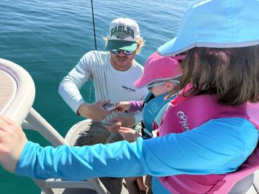 A fishing guide assists two young anglers in handling a fish on a boat, showcasing a fun and educational fishing experience.