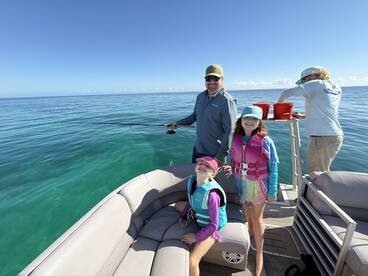 A family enjoying a fishing trip on a pontoon boat, with children wearing life jackets and a parent actively fishing in clear waters.