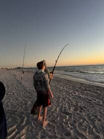 Two young anglers fishing on the beach at sunset, with one actively casting a line into the ocean.
