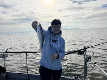 A young angler proudly displaying a striped bass while fishing on a boat, with a scenic water backdrop.
