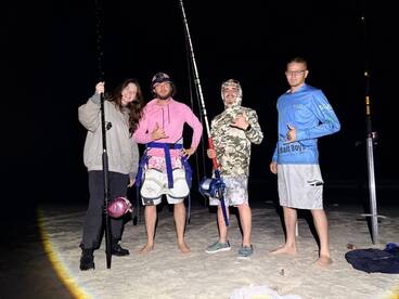 Group of four anglers posing on the beach at night, equipped with fishing rods and gear, showcasing a fun fishing outing.