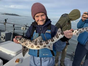 A young angler proudly displaying a Leopard Shark while fishing on a boat, with a scenic backdrop of the bay.