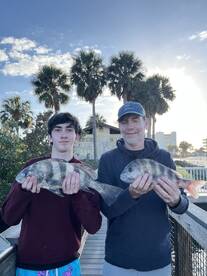 A young angler and an adult proudly displaying their catch of Black Drum fish on a sunny day at the dock.