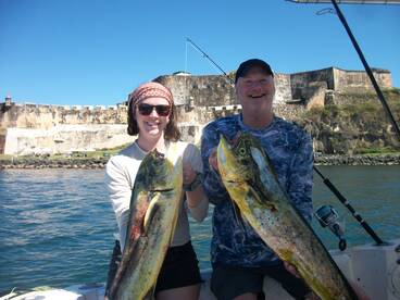 Two anglers proudly displaying their catch of Mahi Mahi in front of a historic fort while enjoying a day on the water.