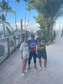 A group of three individuals posing together on a sandy beach, with boats visible in the background, suggesting a fishing trip atmosphere.