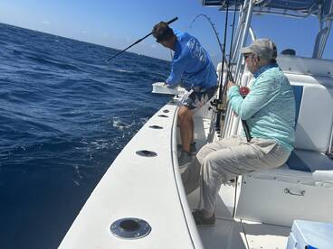 Two anglers actively fishing on a boat, with one reeling in a catch while the other prepares for the next cast in open waters.