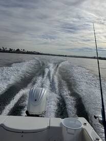 A scenic view from the back of a boat, showcasing the wake and a fishing rod against a cloudy sky, typical of a coastal fishing trip.