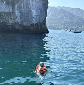 A snorkeler in a life jacket is floating in clear waters near a rocky cliff, with boats visible in the background.