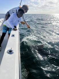 A young angler and an adult are actively engaged in fishing, with a shark visible in the water during a dynamic offshore fishing trip.