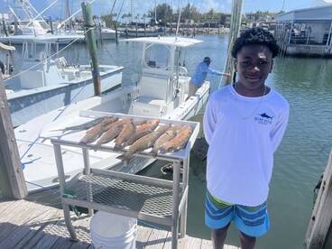 A young angler stands proudly next to a display of freshly caught fish, including Snapper, at the dock after a successful fishing trip.