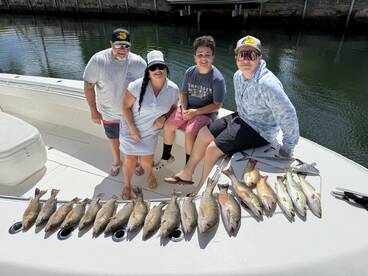 A family poses on a boat with a successful catch of various fish, including redfish and trout, displayed neatly along the side.