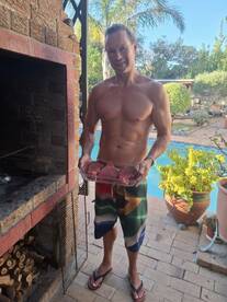 A man holding a tray of fish fillets near a barbecue setup by a pool, showcasing a casual outdoor cooking environment.