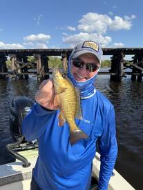 Angler proudly displaying a freshly caught fish against a scenic backdrop of a wooden bridge and clear blue sky.