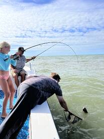 A family enjoys a thrilling moment while fishing on a boat, with one angler reeling in a fish and another preparing to net it.