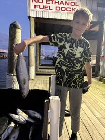 A young angler proudly displaying a large fish at the dock, surrounded by a cooler of additional catches after a successful fishing trip.