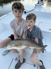 Two young anglers proudly displaying a large Redfish while seated on a boat, showcasing a successful fishing trip.