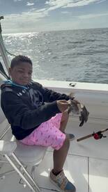 A young angler proudly holding a Black Sea Bass while sitting on a boat, enjoying a day of fishing.