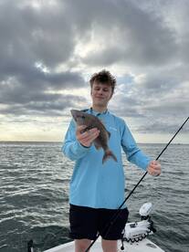 Young angler proudly holding a Snapper while fishing on a boat, showcasing a successful catch against a scenic cloudy backdrop.
