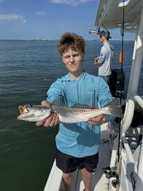 Young angler proudly holding a large Speckled Trout while fishing on a boat in a coastal waterway.