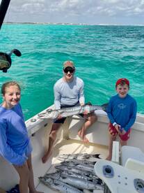 A family enjoying a successful fishing trip, showcasing a large Barracuda and several smaller fish on a boat in clear turquoise waters.