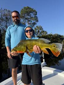 A young angler proudly displaying a vibrant Peacock Bass while fishing with a family member on a sunny day.