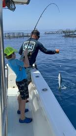 A young angler and an adult are actively fishing on a boat, with a fish jumping out of the water during a vibrant day at sea.