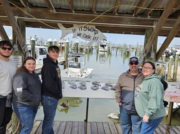 A family poses proudly in front of their catch of flounder displayed on a board at the marina, showcasing a successful fishing trip.