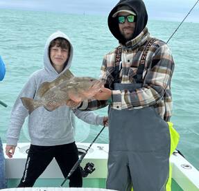 A young angler and an adult proudly display a large Grouper caught while fishing on a boat in coastal waters.