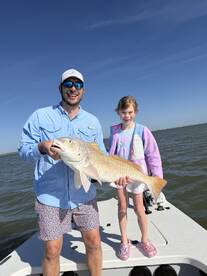 A proud father and daughter duo displaying a large Redfish on a sunny day while fishing from their boat.