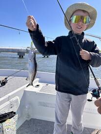 Young angler proudly holding a Speckled Trout while fishing on a boat, showcasing a sunny day on the water.