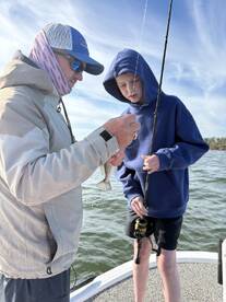 A young angler and an adult are engaged in fishing on a boat, with the adult showing a caught fish, likely a Speckled Trout, to the child.