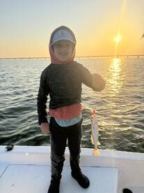 A young angler proudly holding a Speckled Trout against a stunning sunset backdrop while fishing from a boat.