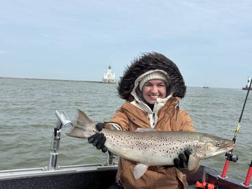 A smiling angler proudly displays a large Brown Trout while fishing on a boat, with a lighthouse visible in the background.