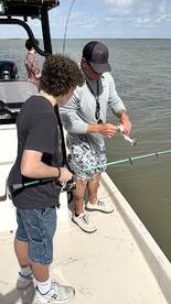 A young angler and an adult are engaged in fishing on a boat, with the adult demonstrating how to handle a small fish caught in the water.