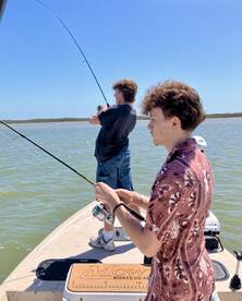 Two young anglers actively fishing on a boat in a scenic waterway, showcasing the excitement of reeling in their catch.