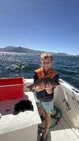 Young angler proudly holding a fish on a boat, with a scenic backdrop of mountains and water during a fishing trip.
