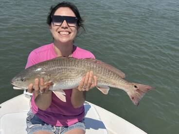 Angler smiling while holding a large Redfish on a sunny day aboard a fishing boat.