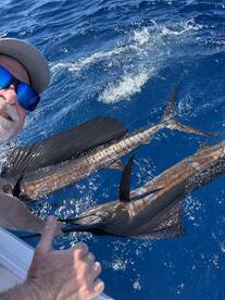 An angler actively reeling in a pair of Sailfish while out on the open ocean, showcasing the excitement of deep-sea fishing.