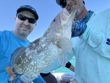 Two anglers proudly displaying a large Grouper while fishing on a sunny day.