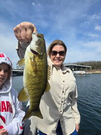 A proud angler displaying a large Largemouth Bass while fishing on a sunny day, with a scenic backdrop of the water and docks.
