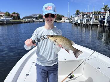 A young angler proudly displaying a large Snapper while fishing from a boat in a scenic marina.