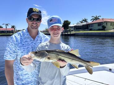 A young angler proudly displays a Snook while fishing with an adult companion in a scenic waterfront setting.
