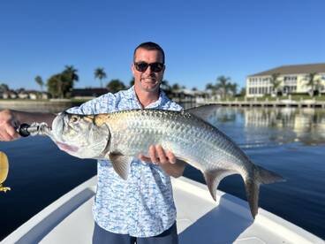 Angler proudly displaying a large Tarpon while fishing from a boat in a scenic waterfront setting.
